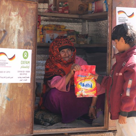 Razigah is sat in the doorway of her small grocery hut handing a red packet to a young boy.