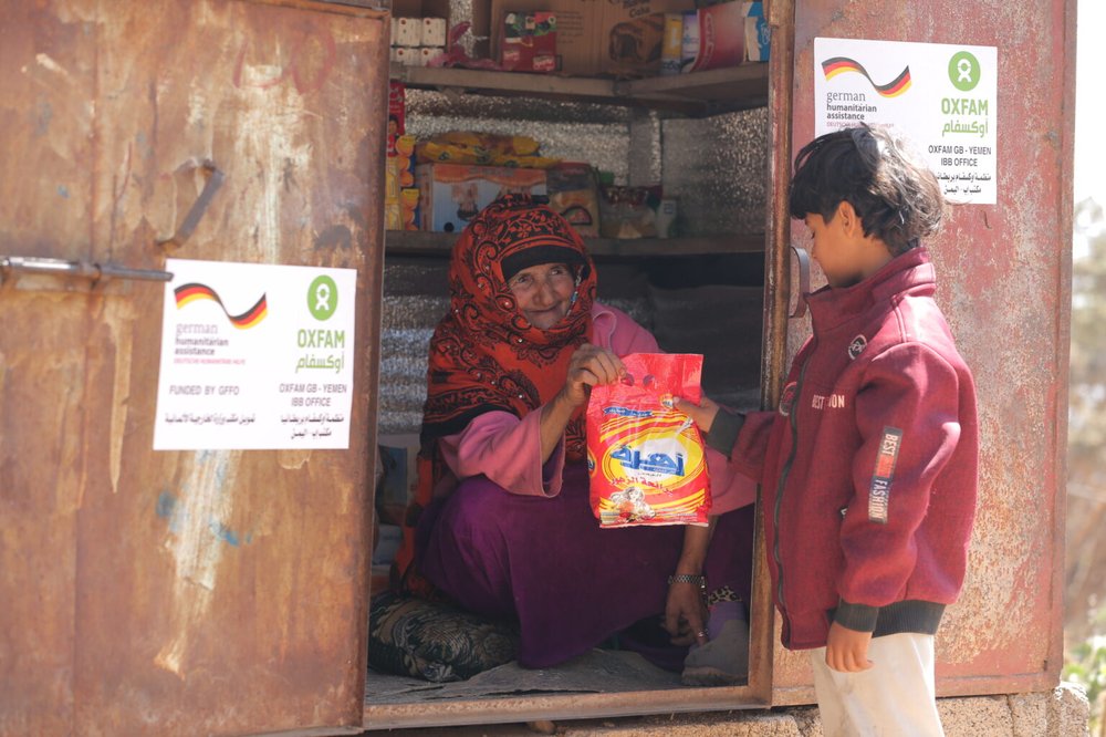 Razigah is sat in the doorway of her small grocery hut handing a red packet to a young boy.