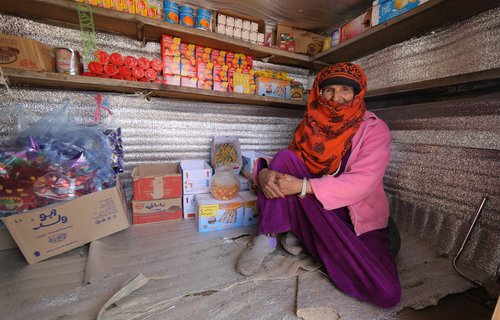 Razigah has a cap and an orange patterened headscarf and smiles with her eyes sitting in her very small metal contanier grocery shop stocked with biscuits and things.