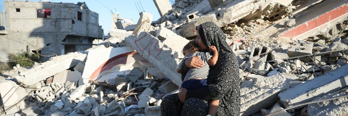 A woman sits amid the rubble of destroyed buildings, holding a small child in her arms. The scene shows devastation and loss in Gaza.
