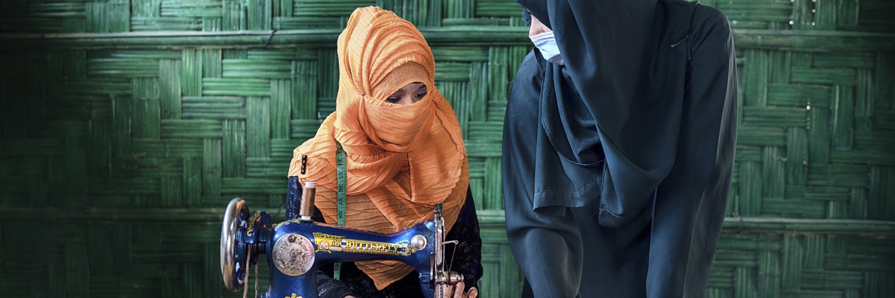 Yeasmin wears a peach coloured hijab that covers her mouth and sits at a sewing machine. The woman standing next to her wears a black hijab and a medical mask.