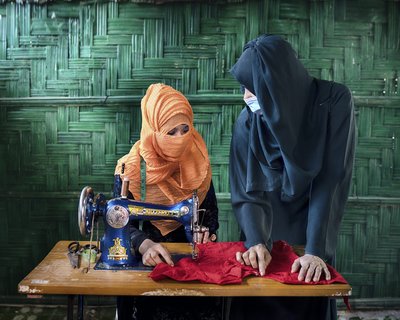 Yeasmin wears a peach coloured hijab that covers her mouth and sits at a sewing machine. The woman standing next to her wears a black hijab and a medical mask.