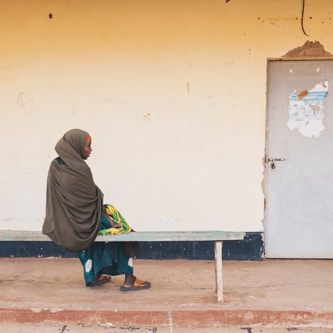 Sowda is wearing brown and organge hijab with blue dress and sandals and sits on a bench outside in Wajir facing a door.