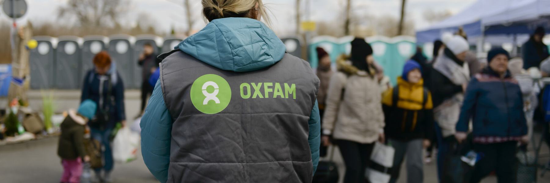The back of a Oxfam staff member with an Oxfam logo on the gilet and people and portaloos in the background.