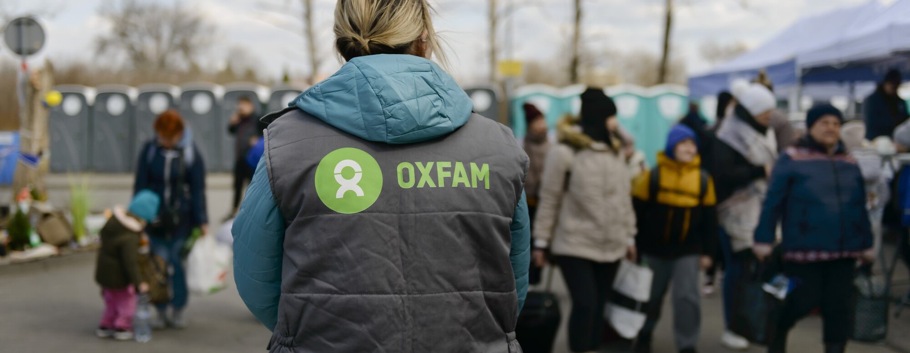 The back of a Oxfam staff member with an Oxfam logo on the gilet and people and portaloos in the background.