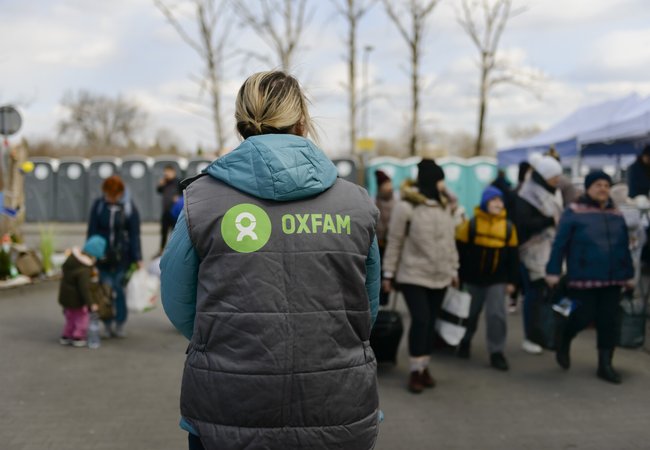 The back of a Oxfam staff member with an Oxfam logo on the gilet and people and portaloos in the background.