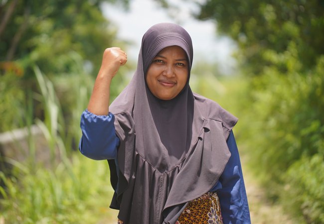 Yuli is an Indonesian woman in her 30s in a light hijab and is raising a fist in an activist pose.