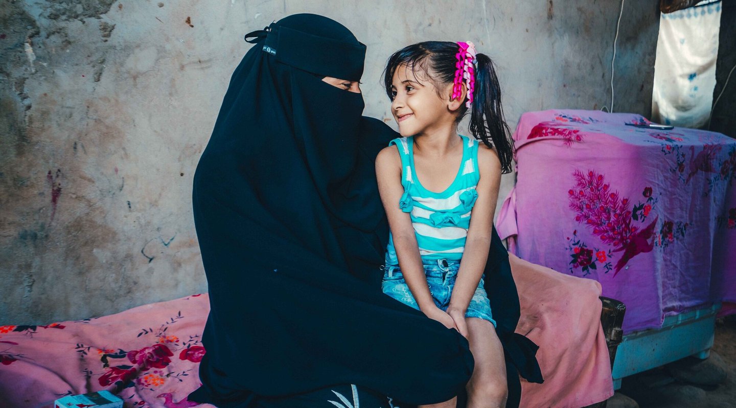 Aryam wearing a black niquab sits with her child, a girl with bunches. They are looking at each other and smiling.