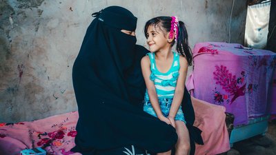 Aryam wearing a black niquab sits with her child, a girl with bunches. They are looking at each other and smiling.