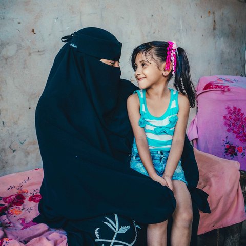 Aryam wearing a black niquab sits with her child, a girl with bunches. They are looking at each other and smiling.