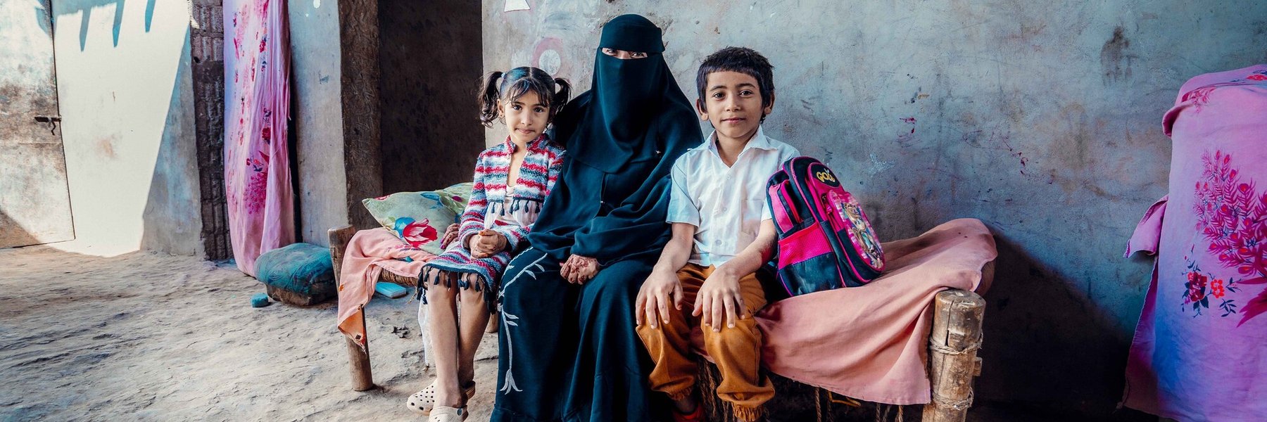 Aryam wearing a black niquab sits with her two children, a girl with bunches and a boy wearing a white school shirt and mustard trousers with a school bag next to him.