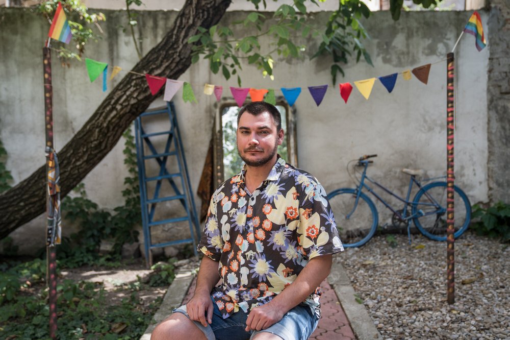 Vlad Levente wearing a bold floral print shirt on a stool in a garden with a blue bike and a mirror against a wall behind him and multi-coloured bunting above him.