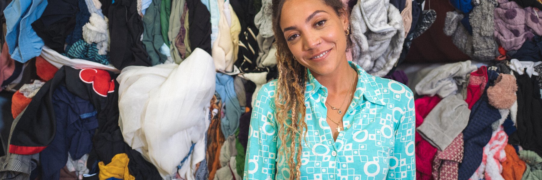 30-something Miquita Oliver has her afro-textured hair brushed back into a ponytail of dip-dyed plaits and curls and smiles warmly. She has a nose ring and wears a light blue shirt and blue jeans. Piles of second hand clothes are behind her.