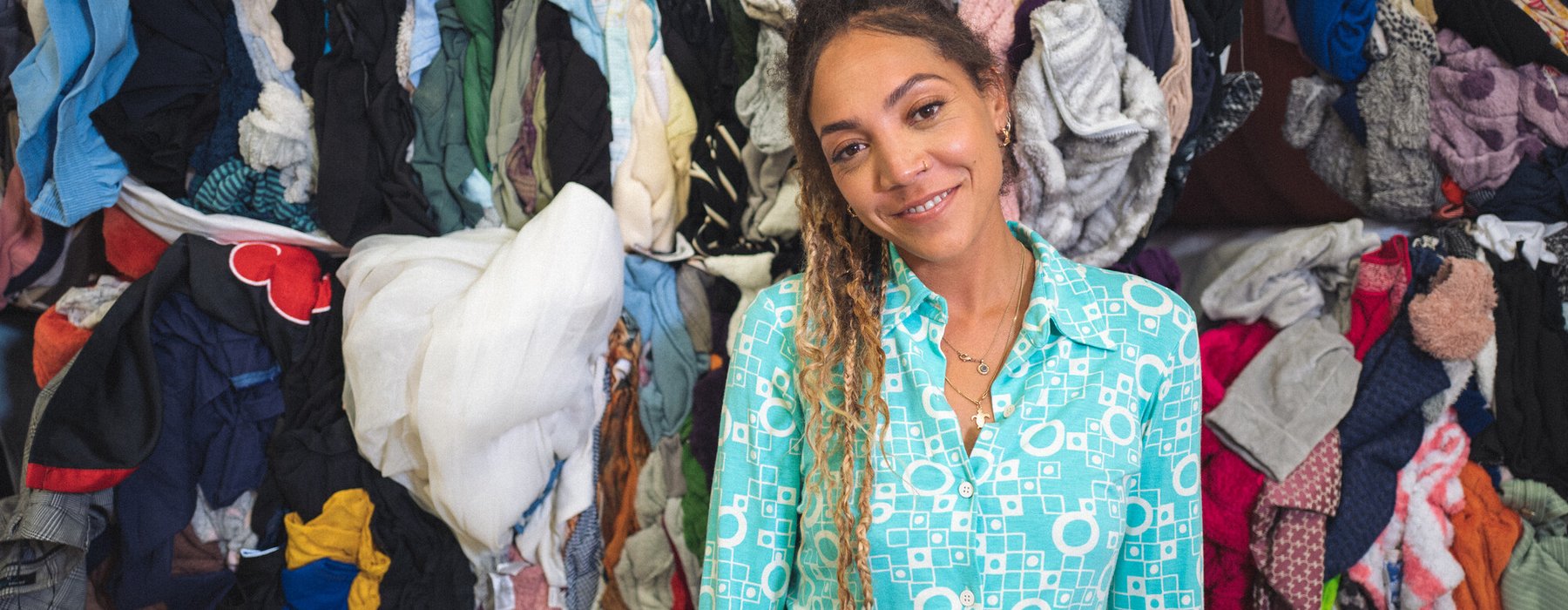 30-something Miquita Oliver has her afro-textured hair brushed back into a ponytail of dip-dyed plaits and curls and smiles warmly. She has a nose ring and wears a light blue shirt and blue jeans. Piles of second hand clothes are behind her.