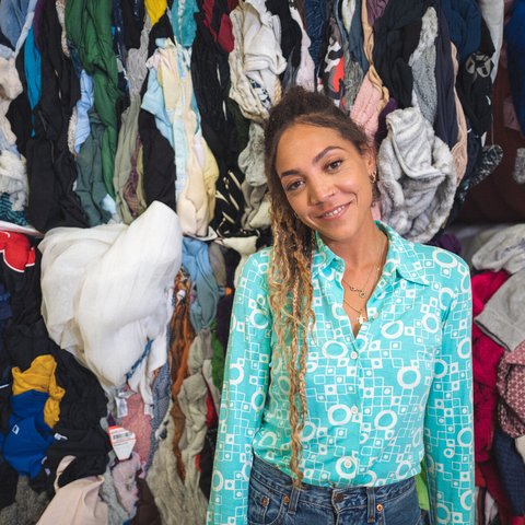 30-something Miquita Oliver has her afro-textured hair brushed back into a ponytail of dip-dyed plaits and curls and smiles warmly. She has a nose ring and wears a light blue shirt and blue jeans. Piles of second hand clothes are behind her.