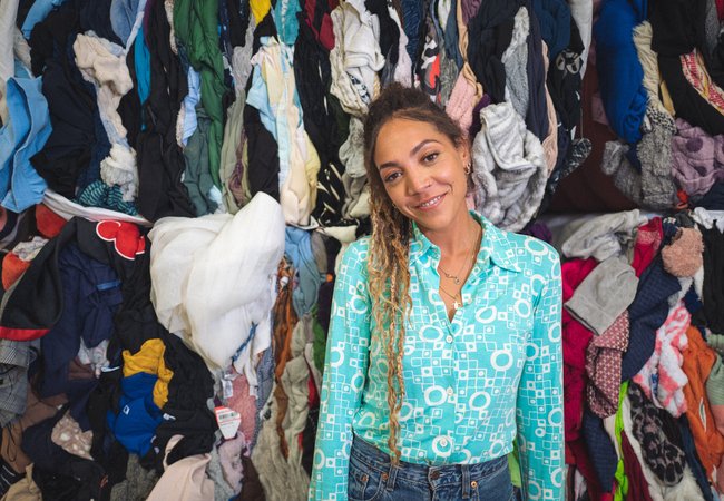 30-something Miquita Oliver has her afro-textured hair brushed back into a ponytail of dip-dyed plaits and curls and smiles warmly. She has a nose ring and wears a light blue shirt and blue jeans. Piles of second hand clothes are behind her.