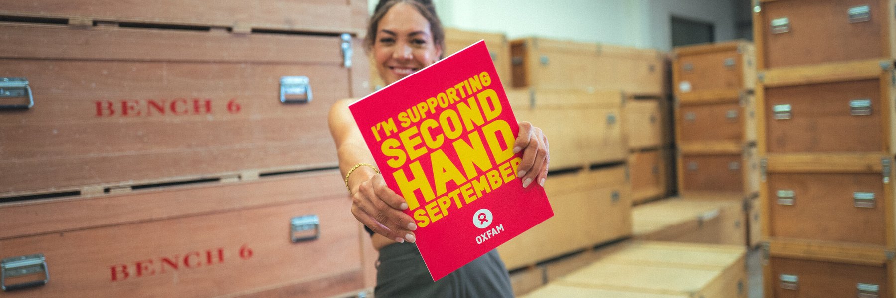 30-something Miquita Oliver has her afro-textured hair brushed back into a ponytail and smiles warmly. She's wearing grey trousers and a crop top and a gold bracelet and holds a sign saying 'I'm taking part in Second Hand Septemer.'