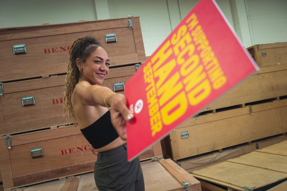 30-something Miquita Oliver has her afro-textured hair brushed back into a ponytail of dip-dyed plaits and curls and smiles warmly. She has a nose ring and wears a crop top and grey trousers. She holds a sign: 'I'm taking part in Second Hand September.'