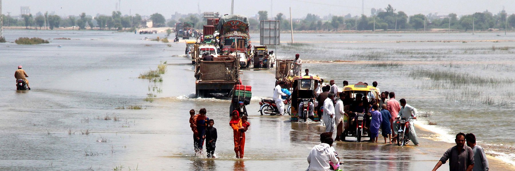 People on foot, on bikes and in vans escape down a flooded highway in Dadu district, Sindh province, Pakistan. Both sides of the road are submerged in flood water, and it covers the road itself.