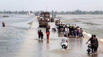 People on foot, on bikes and in vans escape down a flooded highway in Dadu district, Sindh province, Pakistan. Both sides of the road are submerged in flood water, and it covers the road itself.