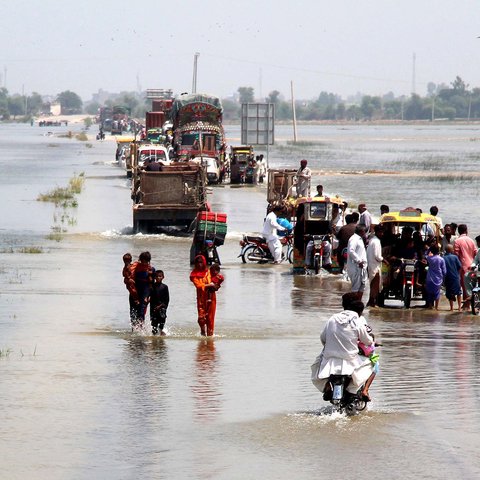 People on foot, on bikes and in vans escape down a flooded highway in Dadu district, Sindh province, Pakistan. Both sides of the road are submerged in flood water, and it covers the road itself.