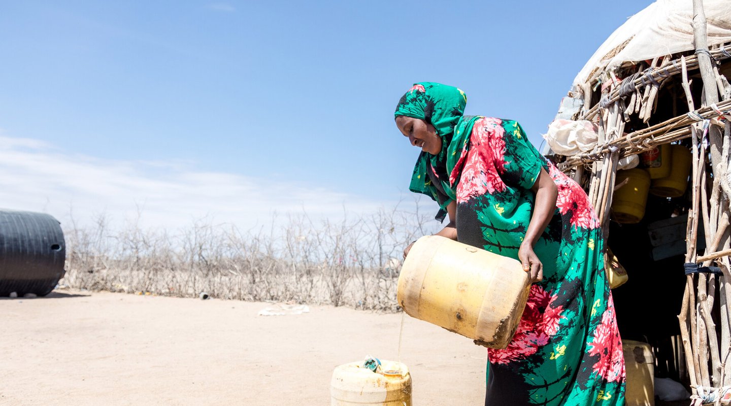 Safia wearing a bright teal and pink hijab and dress collects water in a yellow container in the desert by her home.