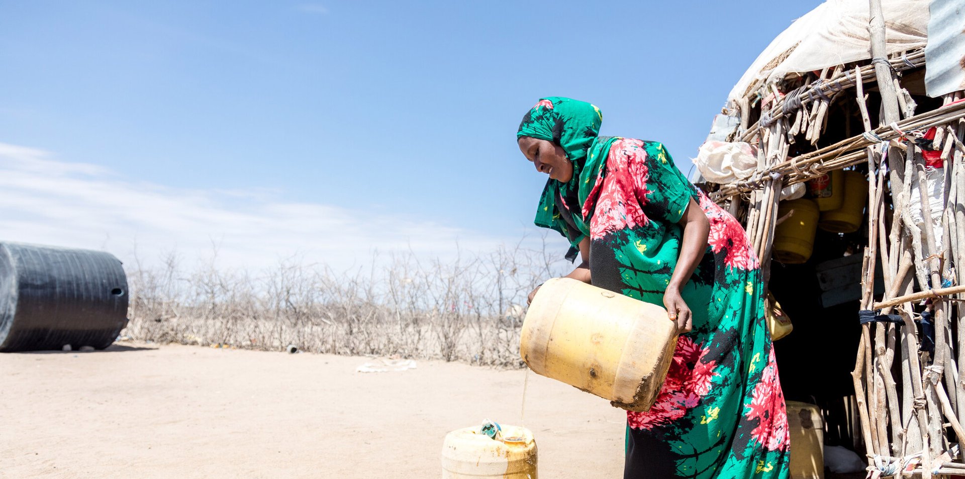 Safia wearing a bright teal and pink hijab and dress collects water in a yellow container in the desert by her home.