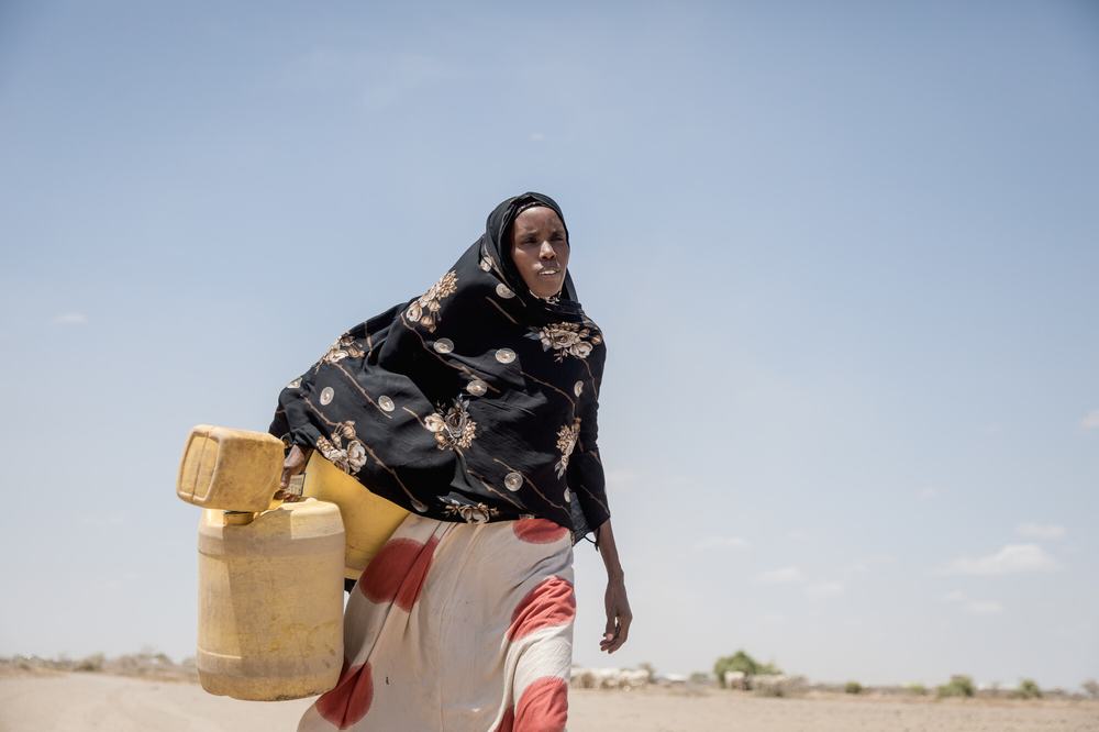 Kenyan Seinhab is in her 30s or 40s and wears a black hijab with gold flowers on it and a whitle long skirt and carries several yellow water containers across the dry land.