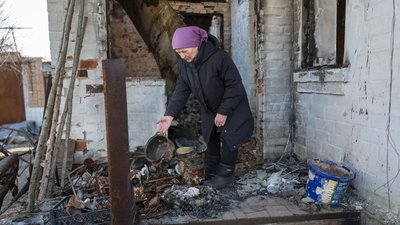 75 year old Nadiya wears a purple scarf on her head and a navy puffer jacket and sorts though fire scorched pots and pans outside her home.