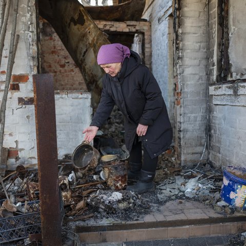 75 year old Nadiya wears a purple scarf on her head and a navy puffer jacket and sorts though fire scorched pots and pans outside her home.