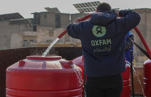 A person wearing an Oxfam jacket is shown with a hose with running water coming out of it into a tank outside in Aleppo.