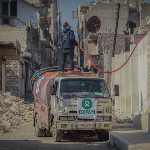 A man wearing an Oxfam jacket stands on a water van with a hose. There is rubble all around.