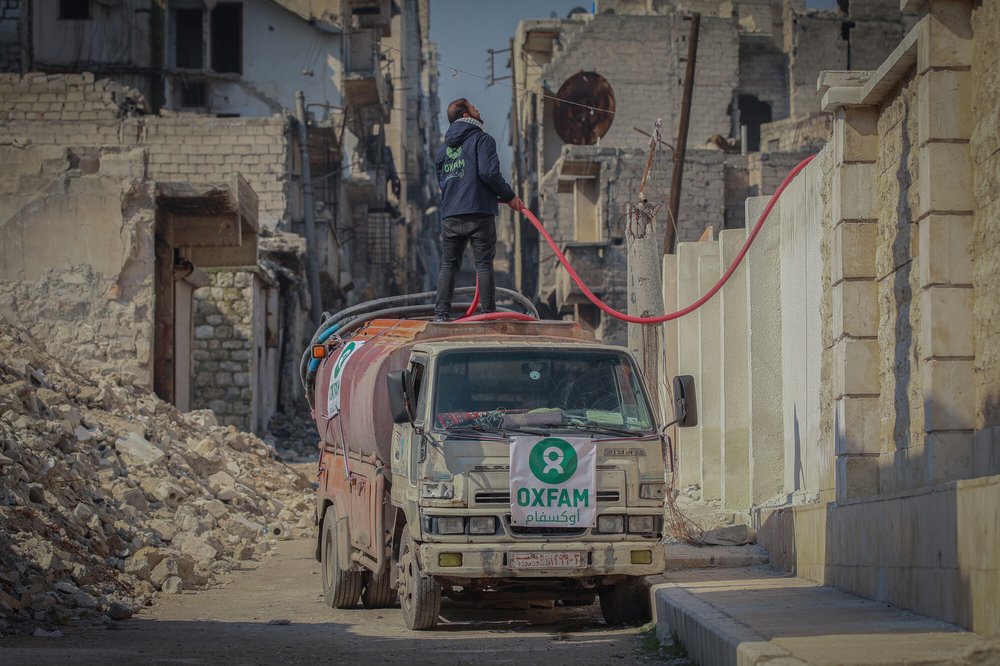 A man wearing an Oxfam jacket stands on a water van with a hose. There is rubble all around.