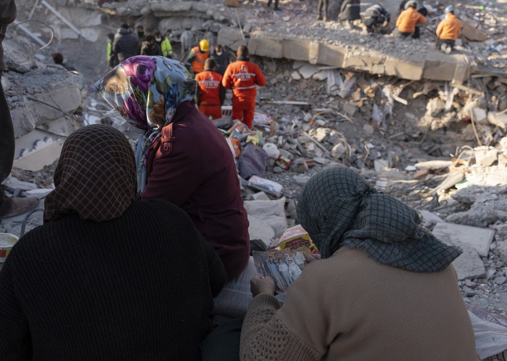 The backs of three women with headscarves, one holds a family photo in her hands. Behind them, rubble and people in orange emergency wear searching among it.