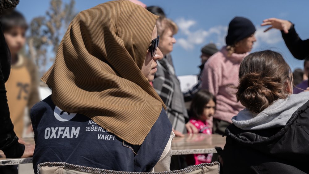 Buthayna stands wearing a brown hijab and sunglasses and an Oxfam vest. There are people in the background.