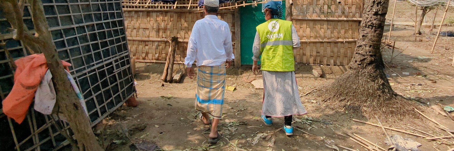 An Oxfam worker stands next to a man in a camp with a hut in front of them with cushions and bags on the roof.