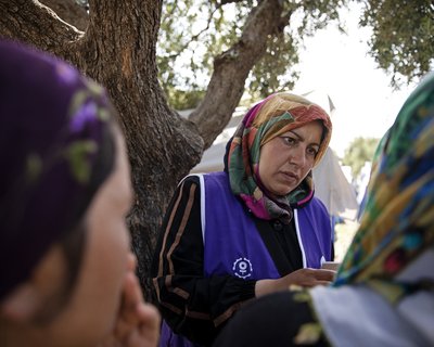 A woman wearing a blue work vest over her abaya and wearing a multi-coloured hijab listens to community members.