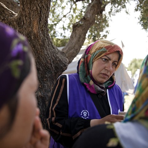A woman wearing a blue work vest over her abaya and wearing a multi-coloured hijab listens to community members.