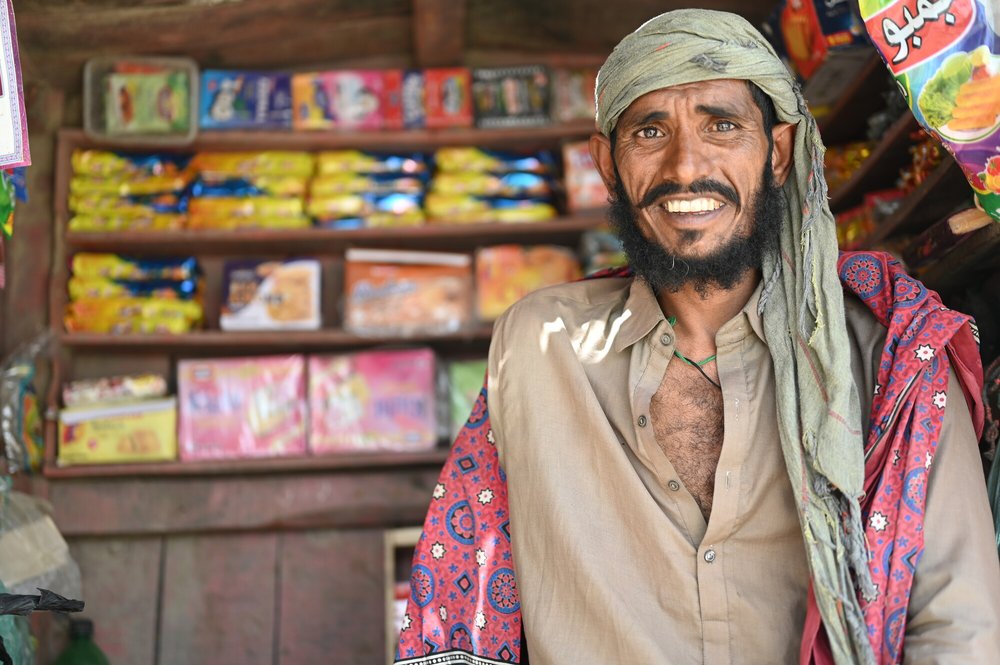 Ghulam smiles in front of his shop with colourful packets of snacks and other foods behind him.