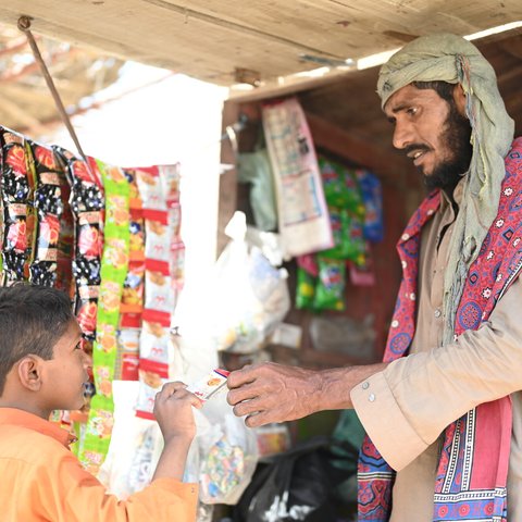 Ghulam hands a card over a counter to a boy.  Ghulam's shop has colourful packets of snacks and other foods.