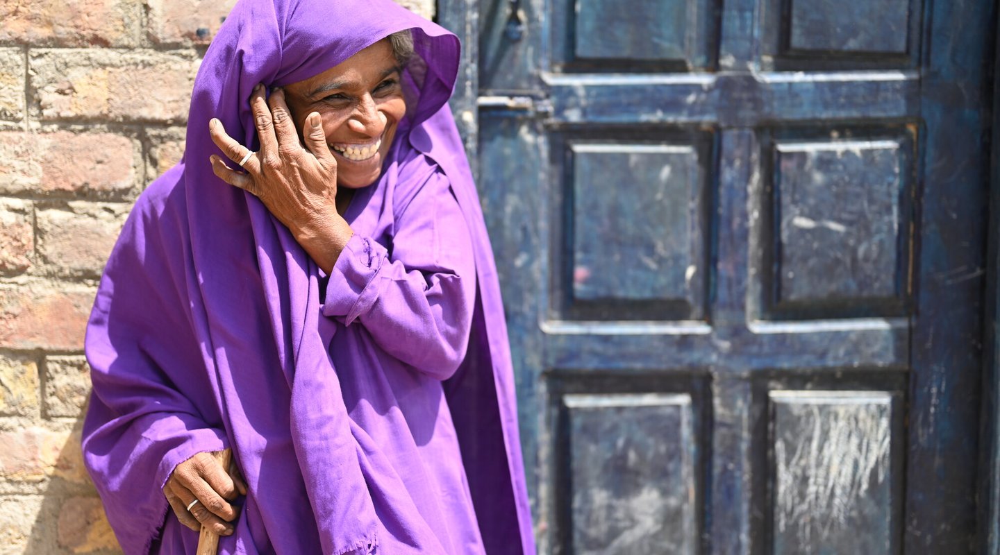 Kisbano wears a purple outfit that covers her hair as well. She smiles and holds her cane and stands outside the village toilet that has just been built.