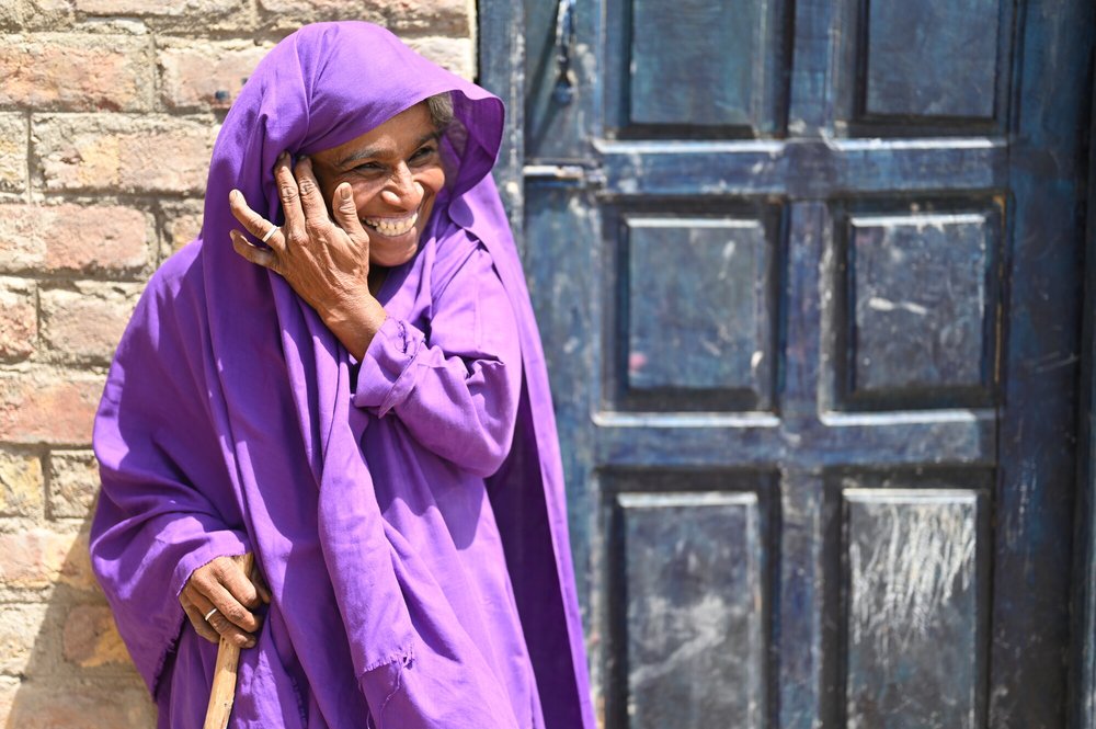 Kisbano wears a purple outfit that covers her hair as well. She smiles and holds her cane and stands outside the village toilet that has just been built.