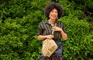 An image of Natalie Lee who has an afro and is wearing a tassel skirt and a tassel bag and a check shirt.