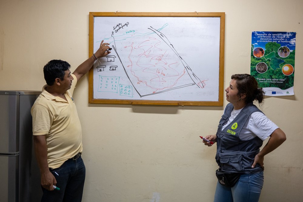 Paul and Paola stand either side of a whiteboard with a drawing of a square of land and red lines hashed over it with green lines around the perimeter.