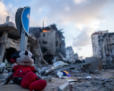 A child's soft toy of Minnie Mouse among the rubble in Gaza and a building in the background on fire.