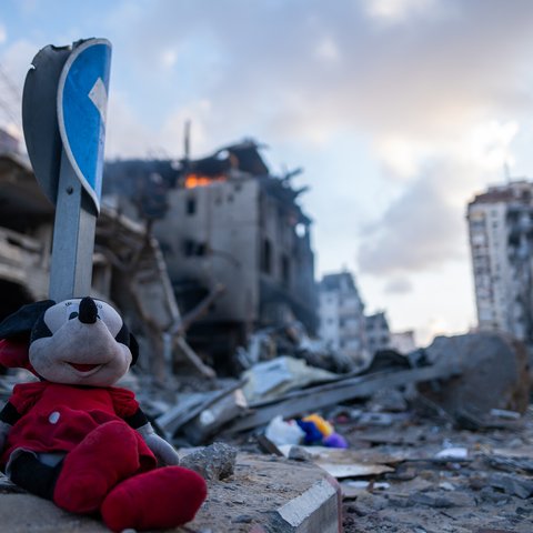 A child's soft toy of Minnie Mouse among the rubble in Gaza and a building in the background on fire.