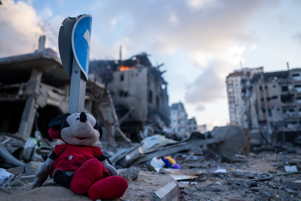 A child's soft toy of Minnie Mouse among the rubble in Gaza and a building in the background on fire.