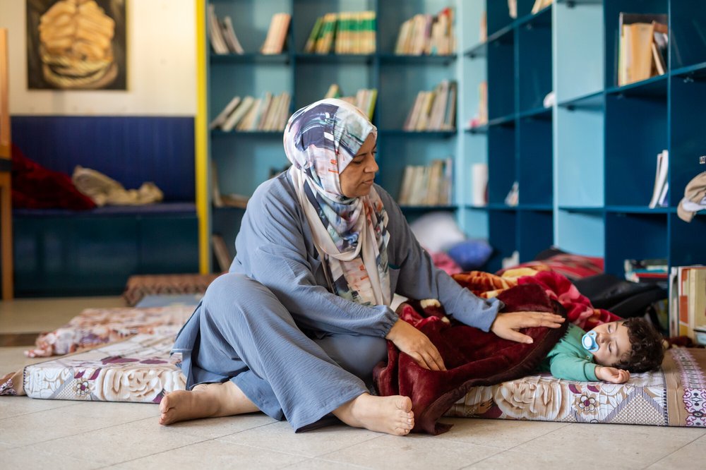 Huwaida wearing a light patterned hijab and grey abaya places a hand gently on a child sleeping on a mat on the floor of a library.