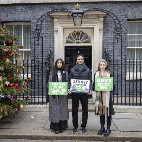 Three people stand by a huge Christmas tree outside Number 10, Downing Street with signs saying that 139,000 people are calling for polluters to pay