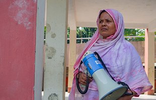 Dulu, a Bangladeshi woman in a pink shawl, stands looking determined while holding a megaphone.